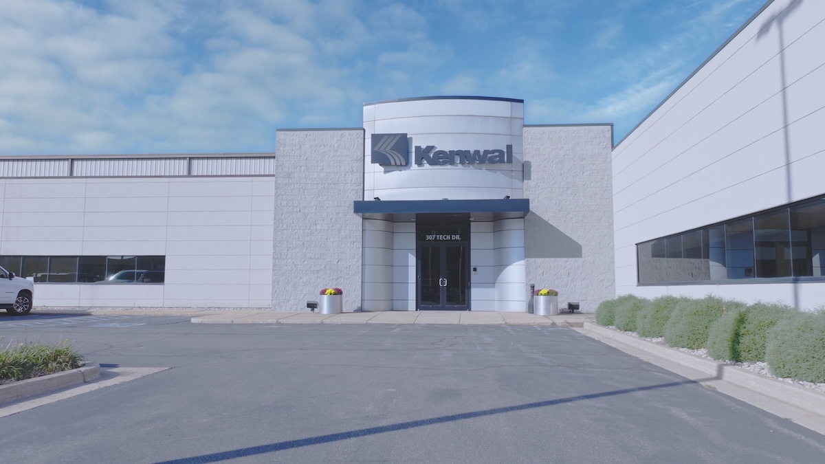 Front entrance of Kenwal Steel corporate office at 307 Tech Drive, featuring modern white exterior, glass doors, company signage, and decorative planters with flowers under a clear blue sky.