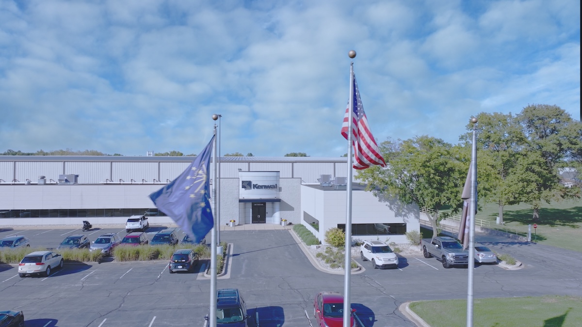 Kenwal Steel Headquarters – Front View with American Flag Front exterior view of Kenwal Steel facility with American flag, Indiana state flag, and company logo on the building facade, surrounded by parked cars and green trees under a blue sky.