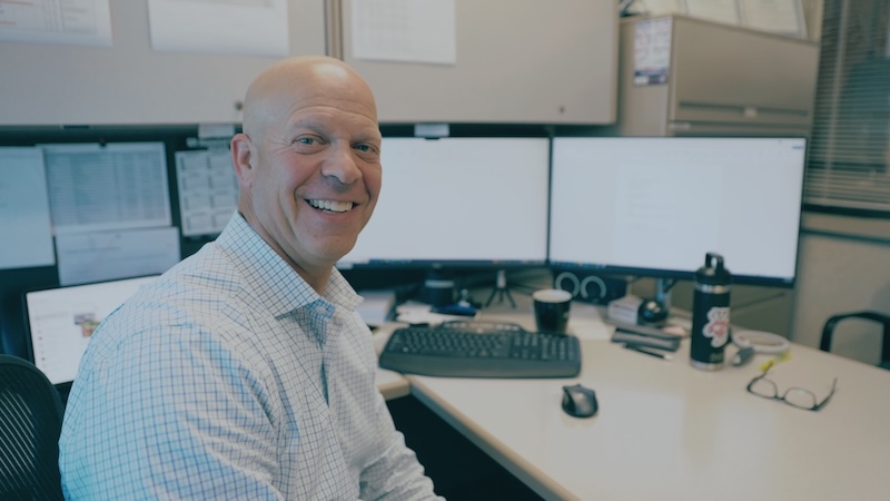 Smiling Kenwal Steel employee working at a dual-monitor workstation in a corporate office setting.