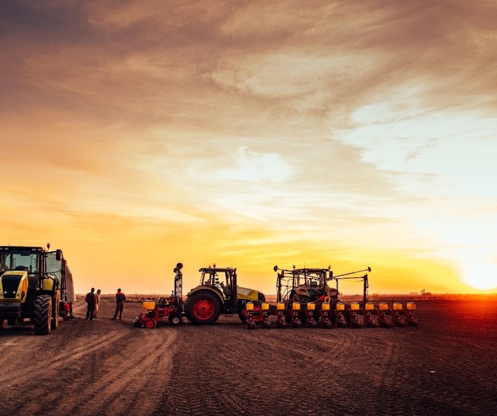 Tractors and advanced agricultural machinery positioned on farmland at sunrise, with workers preparing equipment for farming operations.