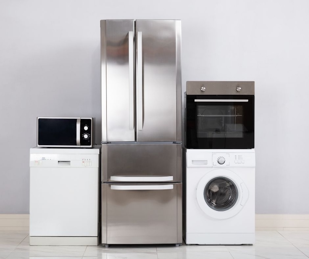 Row of modern kitchen appliances including a stainless steel refrigerator, microwave, dishwasher, oven, and washing machine against a light gray wall.