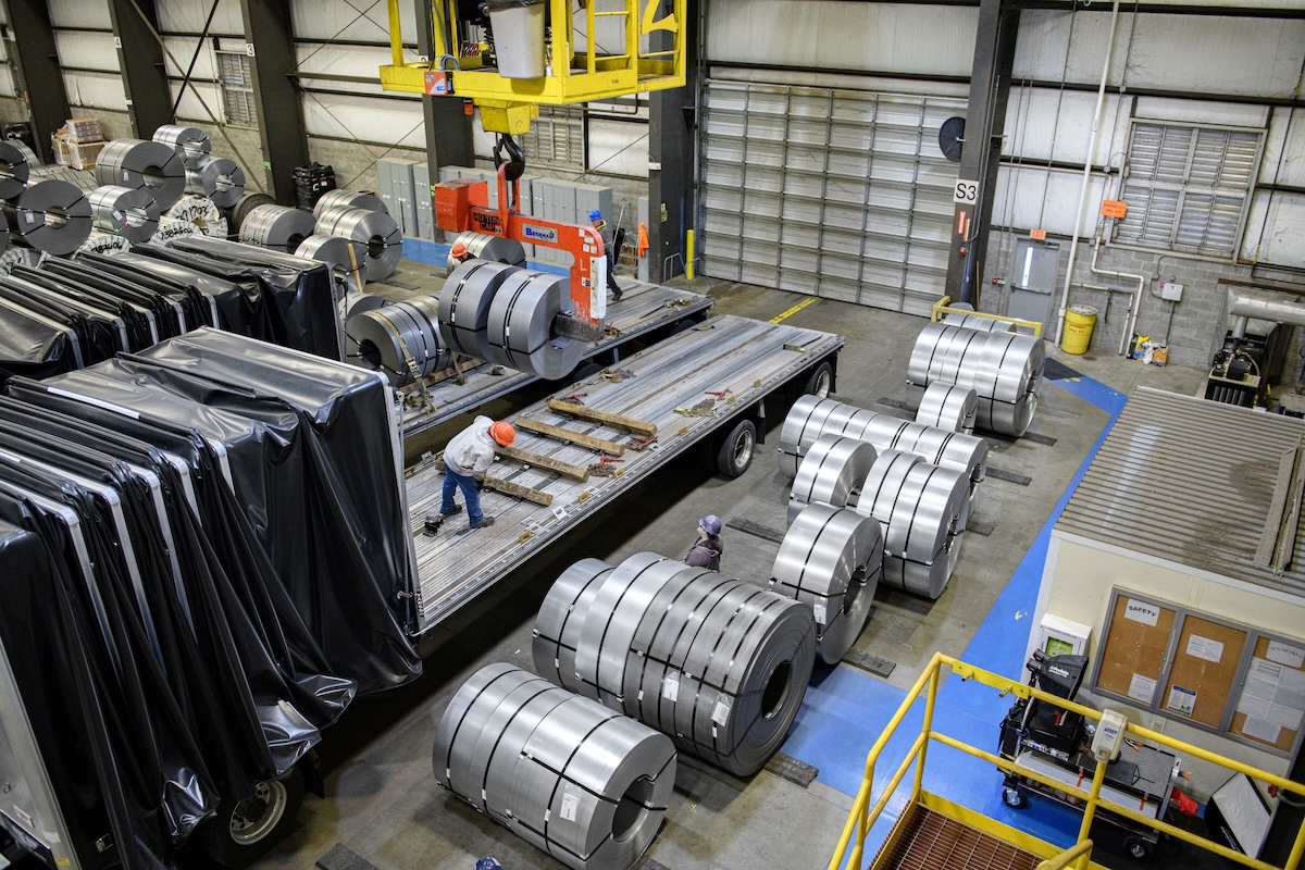 Steel Coil Loading Process | Industrial Shipping and Handling Steel coils being loaded onto a flatbed truck using overhead crane inside industrial warehouse, workers guiding the loading process.