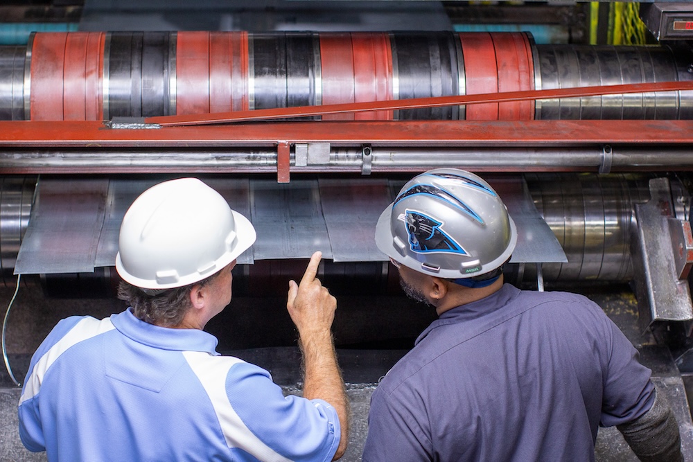 indiana_operations Two steel plant workers wearing hard hats inspecting metal sheets being processed through an industrial roll forming machine.