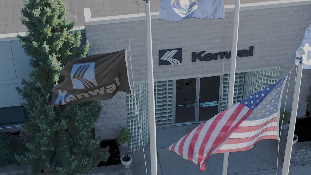 Exterior view of the Kenwal Steel building entrance with company flag, American flag, and Michigan state flag flying in front.