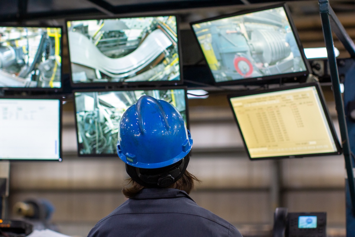Factory operator monitoring multiple production line cameras and control screens at a steel processing facility.