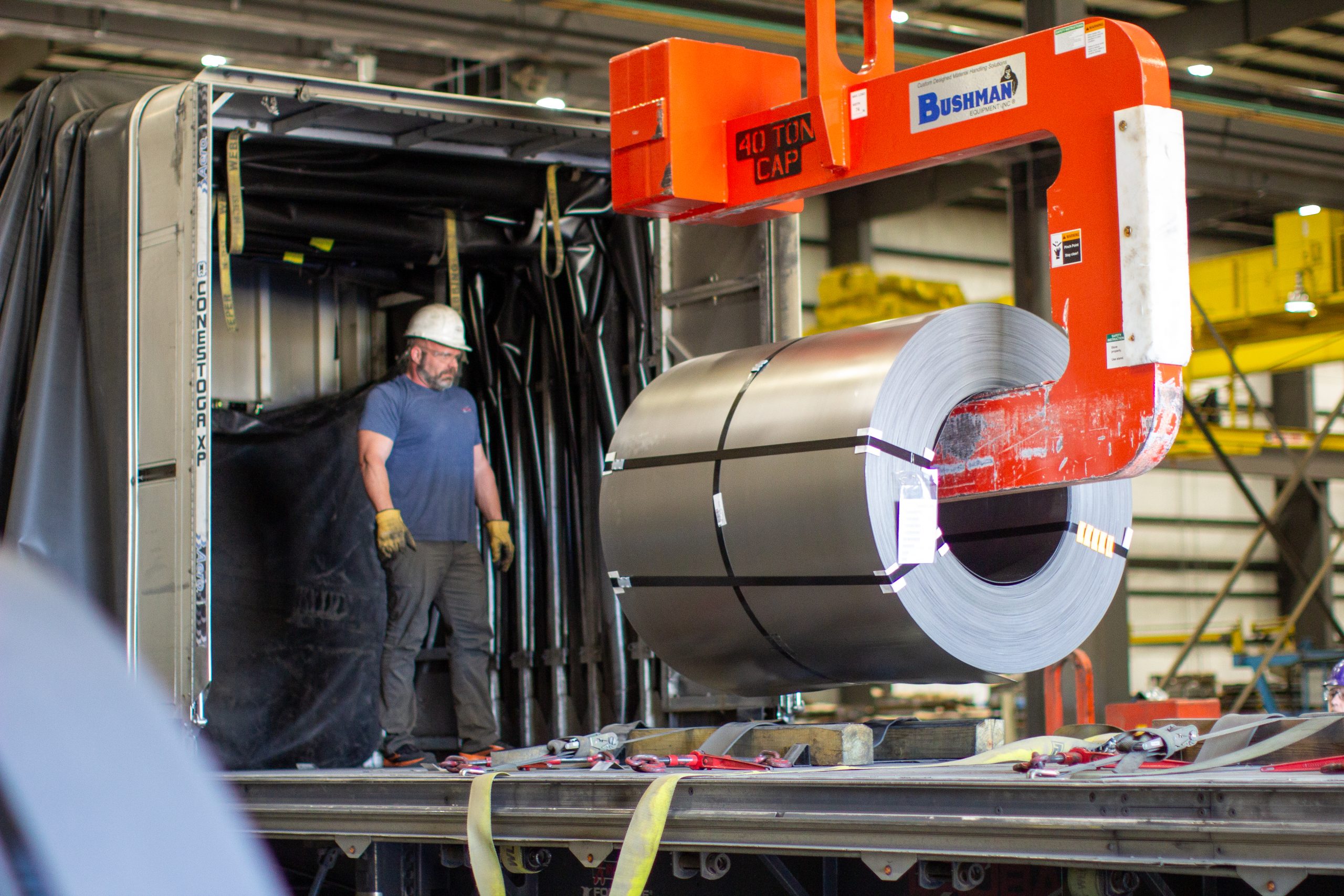 steel-coil-loading-overhead-lifter-logistics Worker overseeing the loading of a steel coil onto a flatbed trailer using a Bushman 40-ton capacity overhead coil lifter in an industrial warehouse.