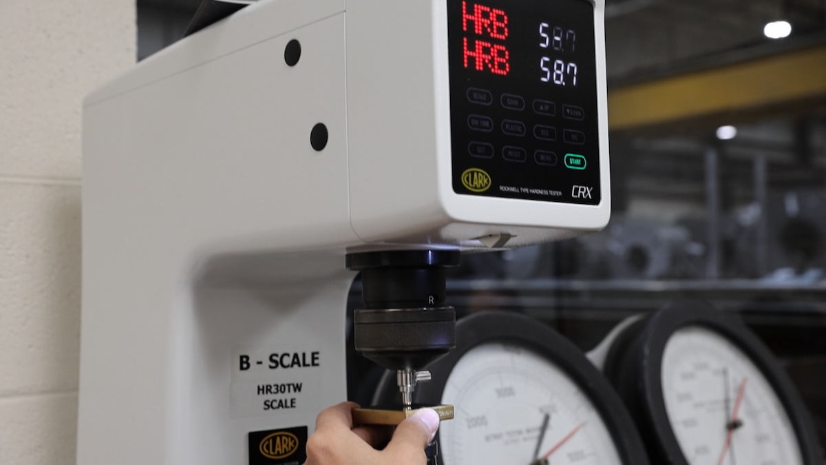 Interior view of a steel service center with stacked steel coils, overhead cranes, and workers operating machinery under high-bay lighting.