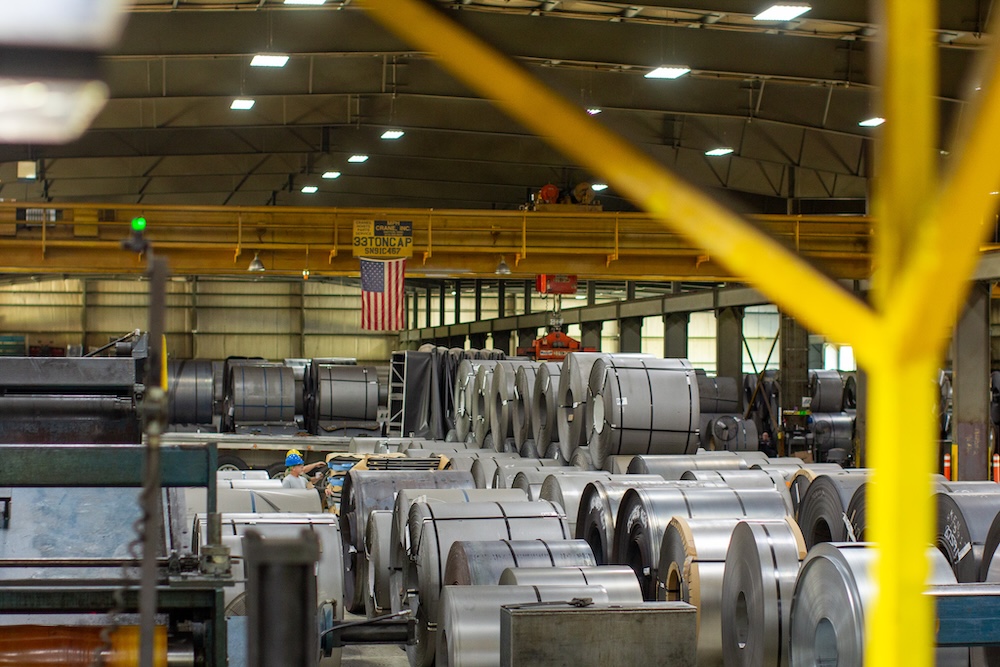 Interior view of a steel service center with stacked steel coils, overhead cranes, and workers operating machinery under high-bay lighting.